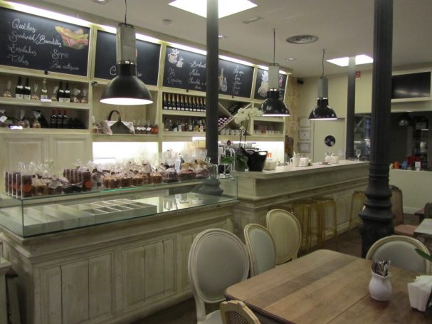 Mostrador de café con dulces, estanterías y mesas de madera en un ambiente acogedor.