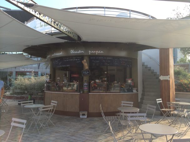 Quiosco moderno con terraza, rodeado de sombrillas y mesas en un entorno al aire libre.